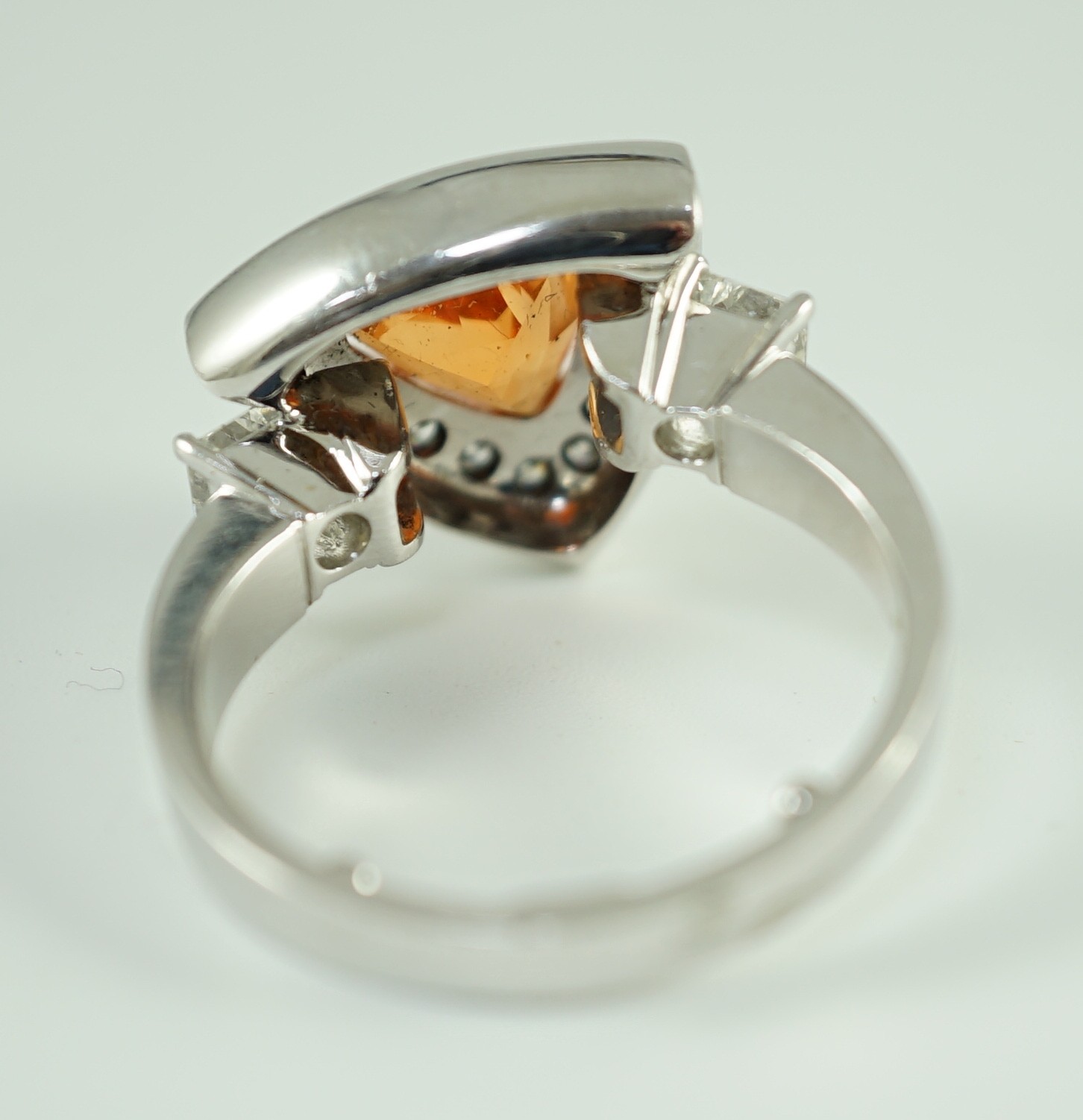 A modern platinum and single stone trilliant cut spessartite garnet ring, with diamond set border and two criss-emerald cut diamond set shoulders, by Musson